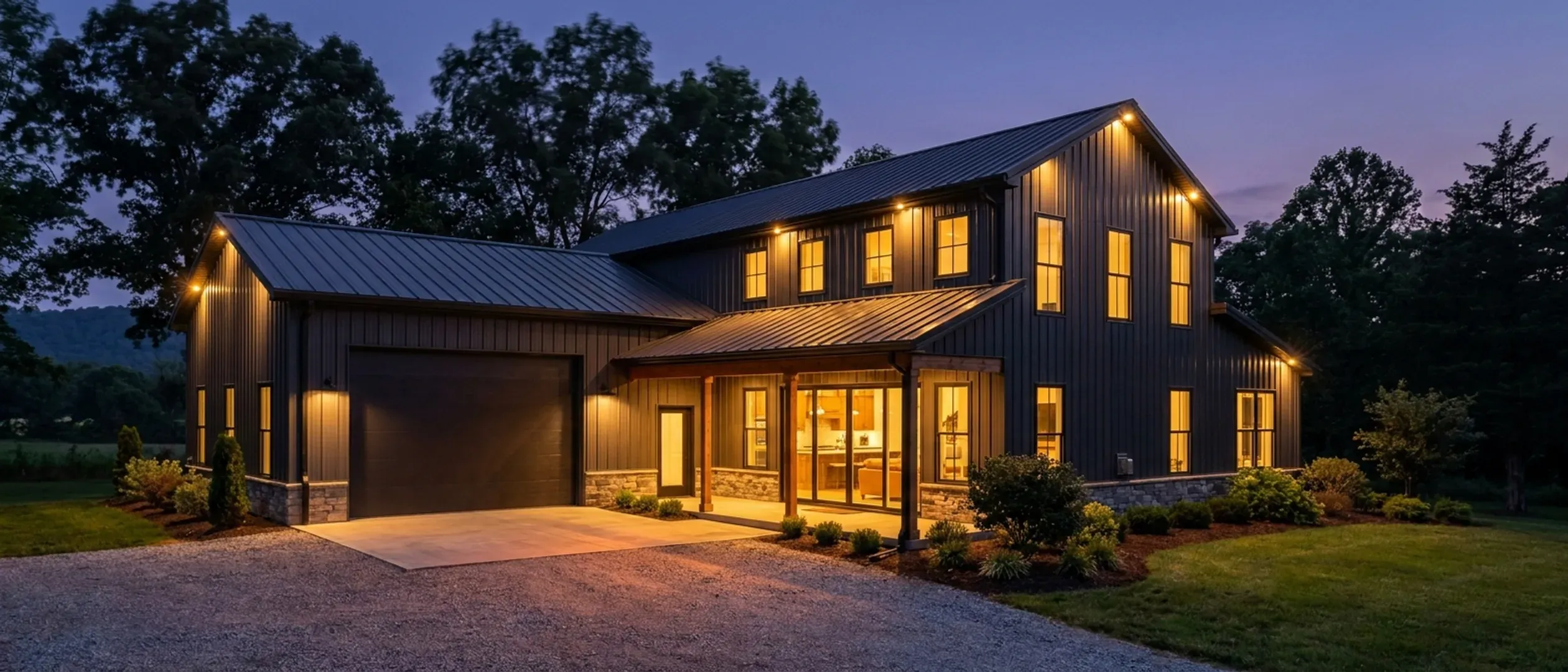 A barndominium at dusk with warm light glowing from every window and exterior lighting along the porch and roofline against a deep blue twilight sky.