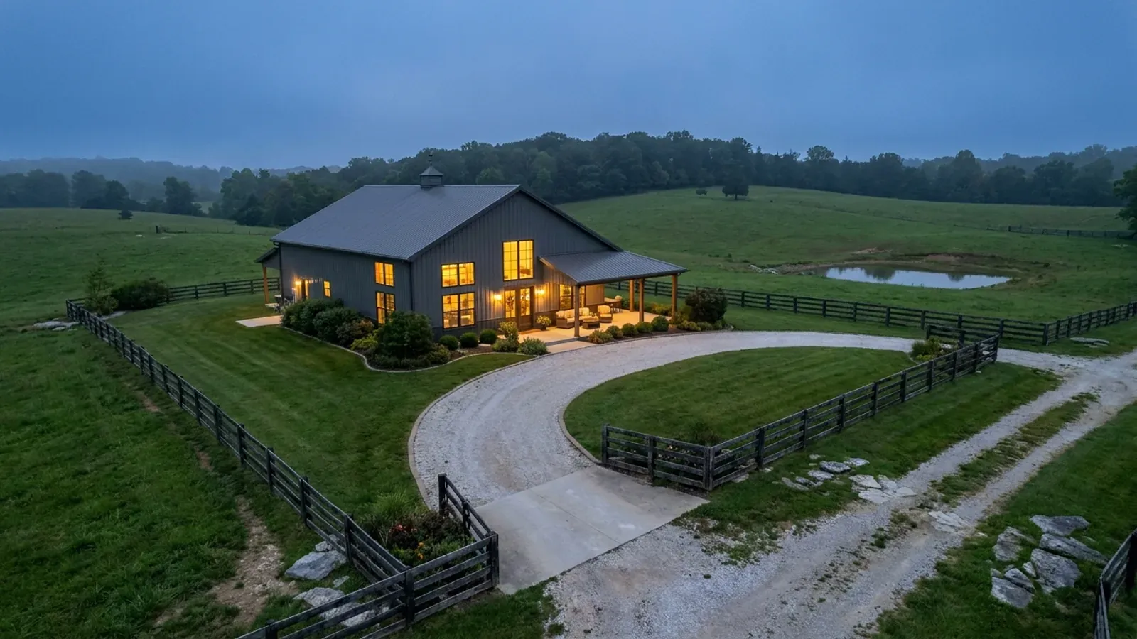 A finished barndominium at dusk with warm interior lighting glowing through its large windows.